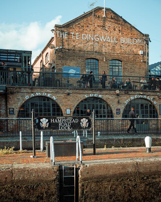A brick building with a sign reading 'The Tingwall Building' is situated along the Camden Lock area, with multiple large arched windows on the ground and upper levels. In front, a black metal railing 