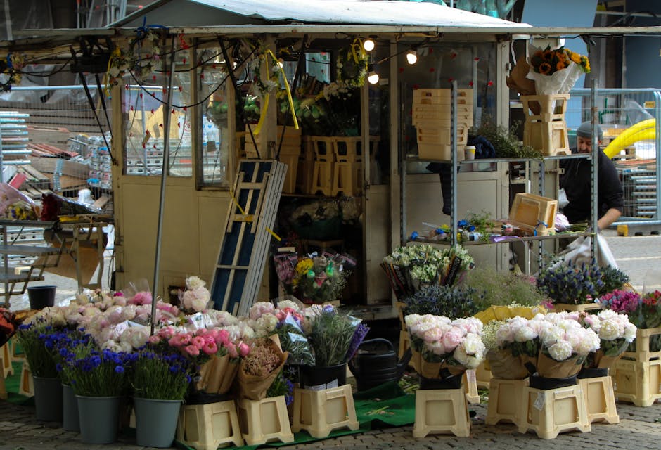 A bustling outdoor flower stall located at Camden Market, displaying an array of fresh floral arrangements. The stall features buckets filled with vibrant pink, white, purple, and blue flowers, includ