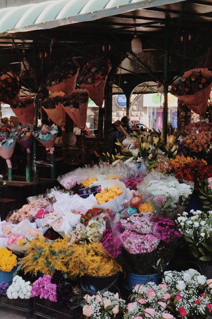 A vibrant outdoor flower stall featuring a diverse array of fresh floral arrangements and bouquets. The display includes large, lush bouquets wrapped in pink, white, and transparent paper, showcasing 