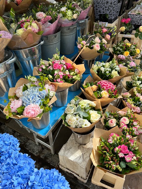 A display of freshly arranged floral bouquets and potted flowers at an outdoor market stall, featuring a variety of pastel-colored roses, peonies, hydrangeas, and lisianthus in shades of pink, white, 
