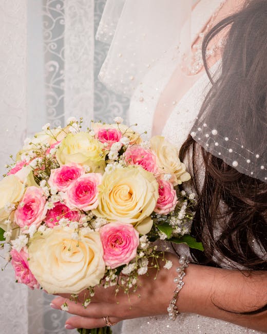 A close-up of a bride holding a floral bouquet featuring large cream roses and smaller pink roses, interspersed with delicate white baby's breath flowers. The bouquet is arranged in a rounded, lush st